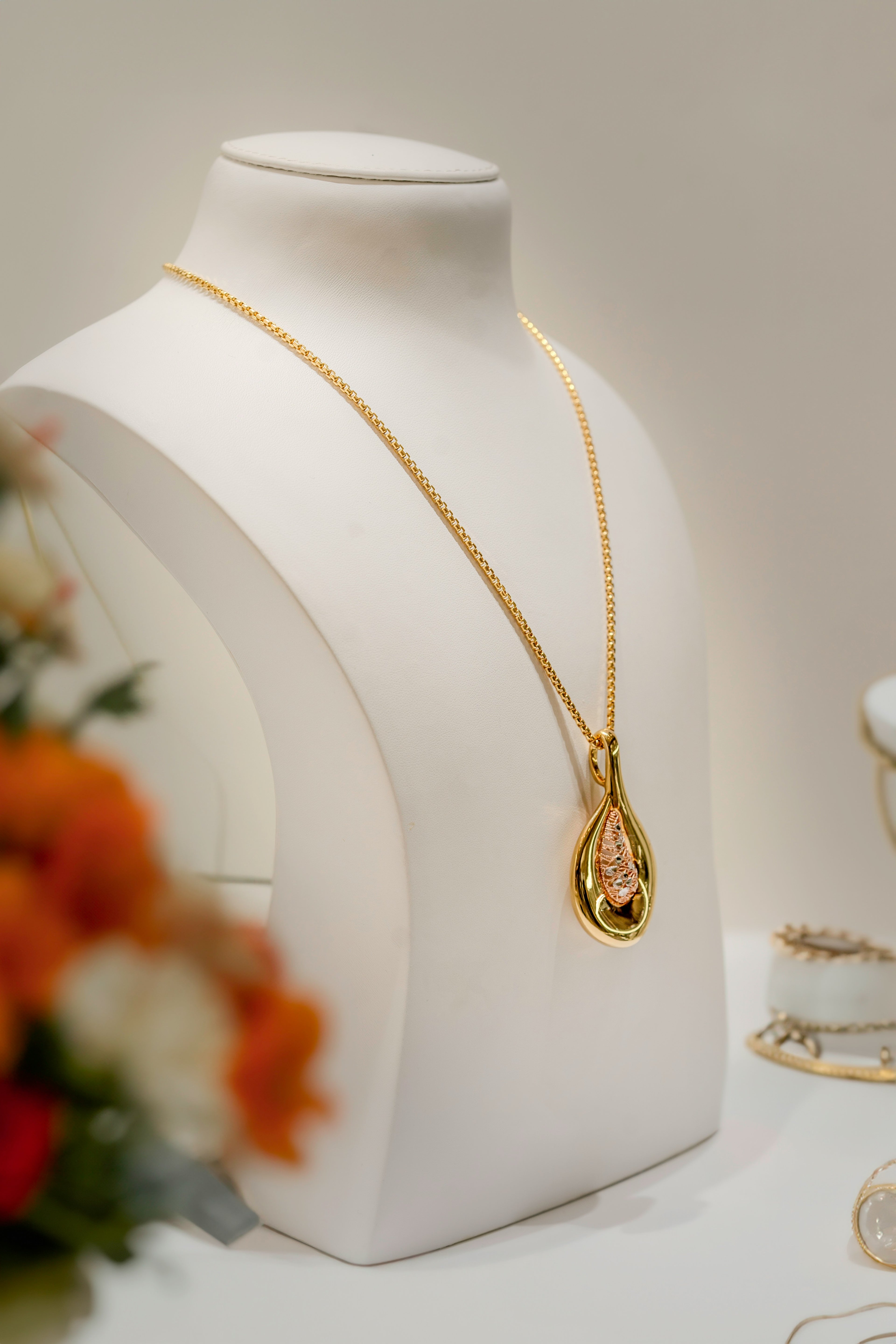 Gold necklace on a mannequin bust with flowers and other jewelry in the background